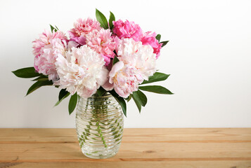 Beautiful bouquet of fresh pink and white peonies in a glass vase sitting on a wooden table against a white wall