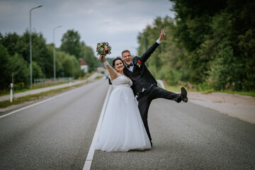 Obraz premium A cheerful bride and groom celebrate on an empty road, the bride holding a bouquet while the groom strikes a playful pose with a peace sign.