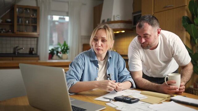 couple sits at table and discusses financial documents in front of laptop. The man points at screen, woman thoughtfully studies finances. Papers, calculator and notepad are scattered on table.