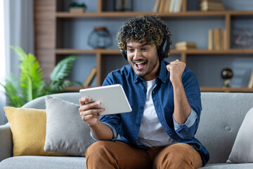 Man sitting with tablet computer at home on sofa, wearing headphones, watching online sports match,...