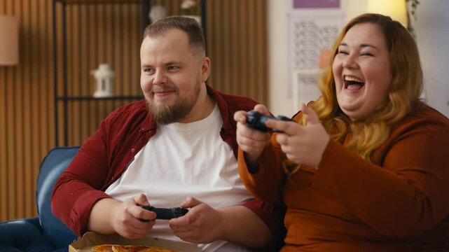 Overweight couple relaxes on the couch, enjoying pizza while playing video games