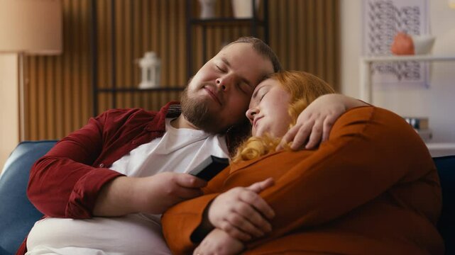 Overweight young couple dozing off on the sofa with the TV on, relaxed lifestyle