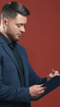 Half-length video of handsome young smiling businessman wearing black shirt and dark blue suit holding the folder taking notes. Dark red background. Vertical video
