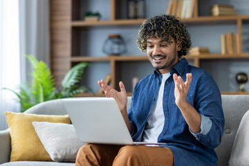 Joyful man sitting on sofa at home, using laptop video call, remote online communication, conference meeting. Businessman working from home in living room. Satisfied student watching video course