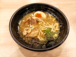 Japanese cuisine. Ramen noodle soup with sliced chicken, egg, chives, sprouts and a piece of seaweed in a black bowl on an isolated wooden table.