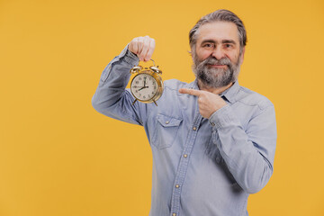 Smiling senior man pointing at alarm clock on yellow background