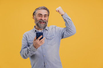 Excited Senior Man Celebrating Success on Smartphone with Raised Fist on Yellow Background