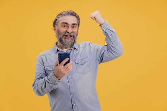 Excited Senior Man Celebrating Success on Smartphone with Raised Fist on Yellow Background - Powered by Adobe