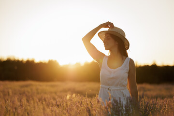 Woman in a lavender field with the sunset in the background. Young woman in white dress and straw hat, smiles and enjoys the end of the day calm and serene.