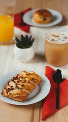 A cozy coffee shop setup featuring two freshly baked pastries, a  cup of coffee, and a orange drink. Photographed at a 45-degree angle. 