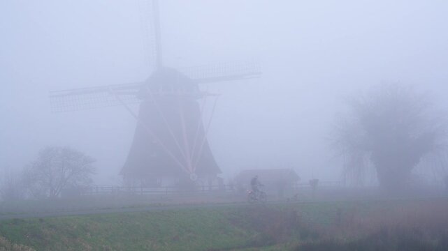 Cycling in The Netherlands in Misty Weather With Low Visibility, Cyclist passes a traditional dutch windmill in a dense fog