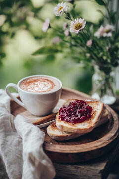 Cozy breakfast with a cup of frothy cappuccino and toasted bread topped with raspberry jam on a rustic wooden tray. Warm, inviting atmosphere with soft natural light and floral background.