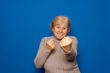 Mature Caucasian woman around 70 years old with her fists in a fighting position against isolated blue background expressing determination. Vitality and self-esteem concept.