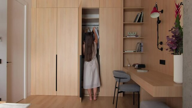 Interior design. Young woman hang clothes in a built-in wardrobe in modern bedroom. Stylish bedroom with cabinet working space bed. Modern furniture at home, storage design