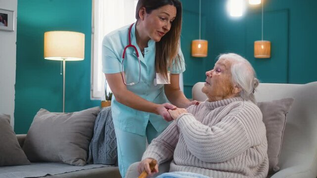 Friendly nurse smiling warmly hugging old 80s woman sitting in armchair, providing compassionate care and helping elderly patients at home