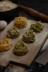 Chopped noodles and rolled into nests lying on a wooden board dusted with flour. Nearby lies a rolling pin, knife, egg and a bowl of flour on a dark table, top view.