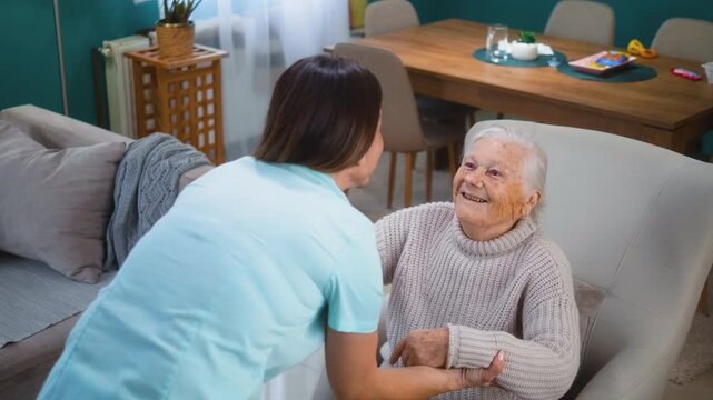 A kind and smiling caregiver in uniform helps an elderly grandmother get up from her armchair at home. The caregiver provides health care and nursing services for elderly people in retirement at home.