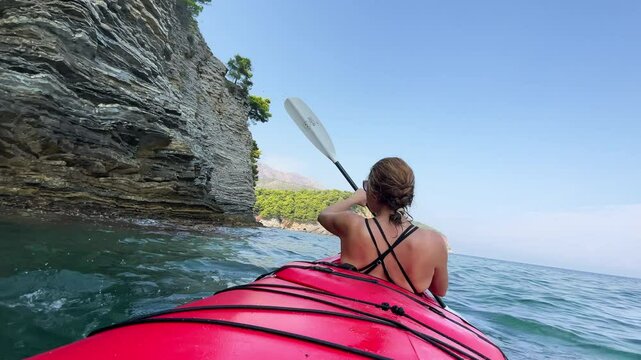 POV 4K video of couple paddling red two-seated kayak along the vertical layered cliff coast by calm Adriatic seashore. Summer holidays in Petrovac, Montenegro.