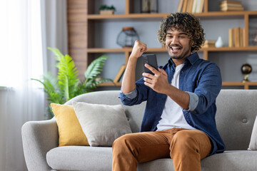 Successful joyful man sitting at home on sofa with phone in hands, celebrating victory and...