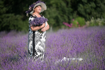 An elegant woman in a black and white dress and wide-brimmed hat stands amidst a vibrant lavender...