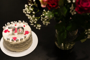 A delicious-looking white cake, decorated with red accents and silver '70' candles, sits on a table next to a bouquet of red roses. A celebratory scene.