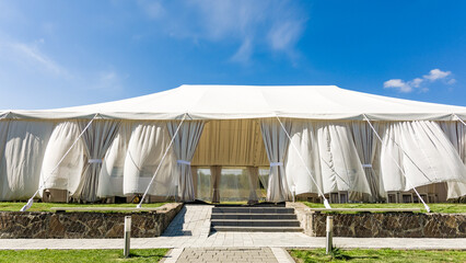 A spacious white event tent stands on a grassy lawn under a bright blue sky.  Curtains adorn its sides, and stone steps lead to its entrance.  Ideal for outdoor gatherings. © Niko_Dali