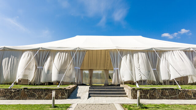 A spacious white event tent stands on a grassy lawn under a bright blue sky.  Curtains adorn its sides, and stone steps lead to its entrance.  Ideal for outdoor gatherings.