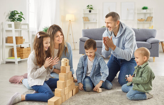 Happy parents playing educational game with their three children, building block toy tower with wooden cubes together on soft carpet in living room. Family activities at home and child creativity. - Powered by Adobe