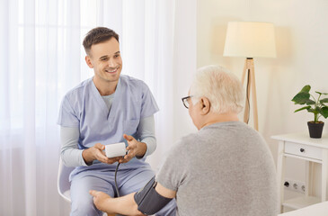 Smiling caregiver measuring blood pressure of senior patient sitting in clinic or nursing home...