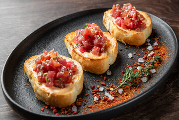 Three toasted bread slices topped with fresh tomato salsa and herbs, served on a black plate. Garnished with rock salt and red pepper flakes, creating a vibrant and appetizing presentation.