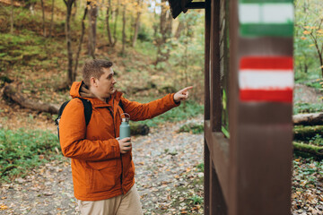 Obraz premium Happy traveler man pointing on wooden billboard with map in the nature. Portrait of young handsome man with backpack wanderer walk in the woods while enjoying the nature landscape. High quality photo