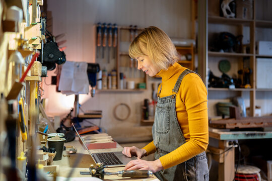 Skilled female artisan works diligently on commissions and order bills on laptop in woodwork workshop. Supply chain, process bills, handle invoice and tackles. Joiner manager responsibility at work