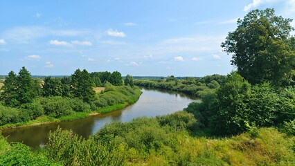 Fototapeta premium The river makes a turn among grassy banks with shrubs and deciduous trees. The trees and blue sky are reflected in the calm water. Meadows are spread out on the floodplain and the forest beyond. Sunny