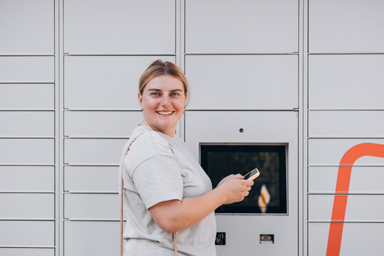 Parcel delivery machine. Mail delivery and post service concept. Happy smiling woman with phone at outdoor automated parcel machine. Electronic locker for storing parcels.