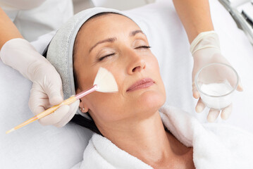 Cosmetologist applies cosmetic product on middle aged woman face for moisturize face skin in beauty...