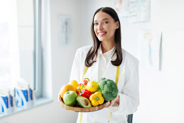 Portrait of European woman nutritionist in office posing with plate of fresh fruits and vegetables,...