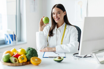 European woman dietitian holding apple, sitting at desk with plate of fruits and vegetables,...