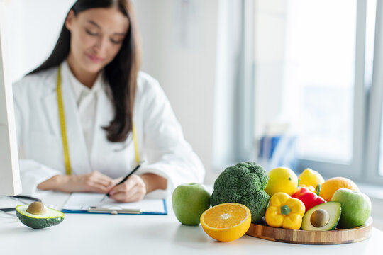 Health and beauty. Professional dietician doctor signing diet plan, working at desk with focus on fresh fruits and vegetables