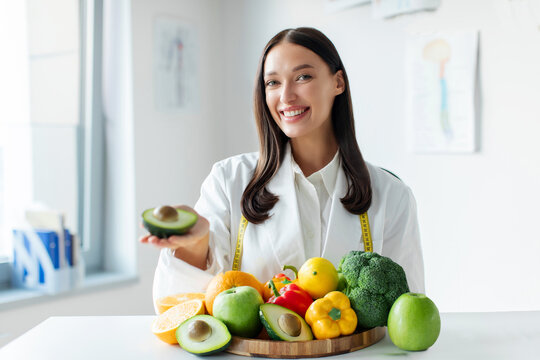 Positive European woman dietologist holding avocado, recommending fresh fruits and vegetables for diet, sitting at desk in medical office and smiling at camera