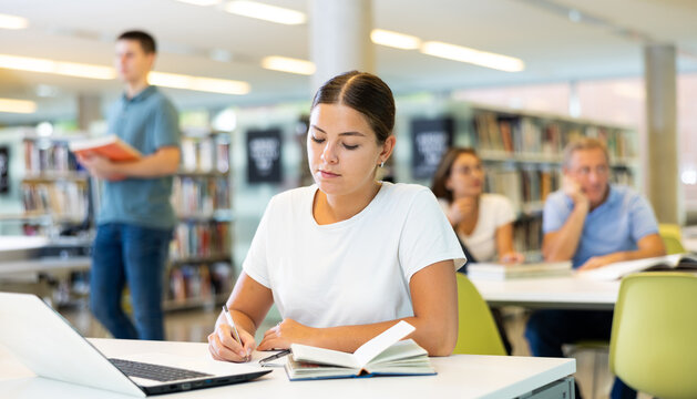 Portrait of positive woman with laptop and book in public library