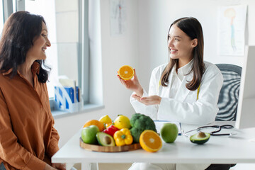 Nutritionist woman giving consultation to mature female patient, holding half of orange and talking...