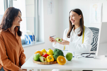 Professional nutritionist talking to mature female patient about benefits of healthy diet while holding avocado, discussing treatment plan for gastrointestinal tract sitting in clinic office
