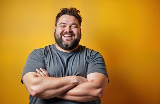 Cheerful man with beard stands confidently against mustard yellow background. Wears casual gray t-shirt, arms crossed. Expression joyful, happy. Portrait positive body image, celebrates