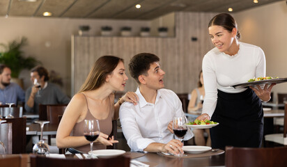 Friendly waitress bringing dishes salad to guests of cozy restaurant