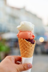 A hand holding a delightful ice cream cone featuring three colorful scoops of ice cream in white, pink, and purple. The cone is a crispy waffle cone, perfect for a hot summer day.