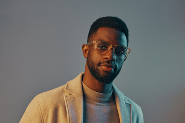 Stylish African man in glasses, wearing a light sweater and blazer, poses confidently against a gradient background, showcasing a contemporary fashion look
