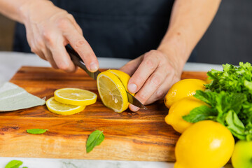 Close-up of hands slicing fresh lemons on a wooden cutting board with a bundle of whole lemons, mint leaves, and a green towel on a marble surface.