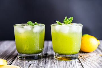 A refreshing green lemonade served in a glass with ice, garnished with mint and lemon slices, placed on a wooden board with fresh lemons and herbs in the background.
