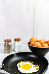 A sunny-side-up egg in a nonstick pan on a textured surface, surrounded by a spatula, bowl of eggs, and salt and pepper shakers for a classic breakfast vibe.