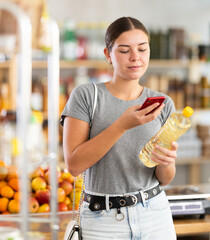 Smiling woman scanning a bottle of vegetable oil through an online app on mobile phone. Visitor...